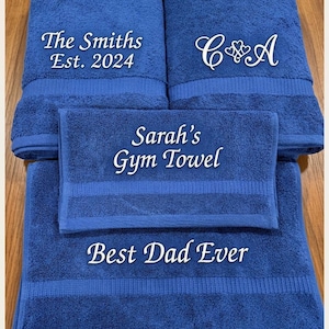 May include: Four folded royal blue towels featuring white embroidered text. The top left towel reads The Smiths Est. 2024, the top right features C and A with hearts, the centre towel reads Sarah's Gym Towel, and the bottom towel reads Best Dad Ever.