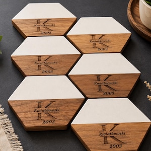 May include: A set of six hexagonal coasters featuring a two-tone design with a white top section and a natural wood bottom section. Each coaster is engraved with a large letter K, the name Kwiatkowski, and the year 2003 in a dark font.