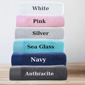 May include: A stack of six plush bath towels in various colors: white, pink, silver, sea glass, navy, and anthracite. Each towel has a contrasting border and a label with its color name.