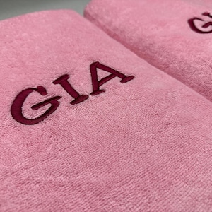May include: Two soft, light pink towels featuring the name GIA embroidered in a dark magenta thread. The fabric has a plush, textured surface, and the lettering is clearly visible on the front of each folded towel, showcasing a clean design.