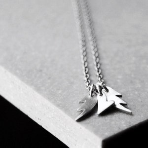 Puede incluir: Un collar de plata con tres colgantes: un corazón, un rayo y una lágrima. El collar está sobre una superficie gris.