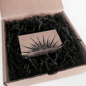May include: A small cardboard box with black shredded paper inside. The box has a brown paper label with a sunburst design and the text "SOU" printed on it.