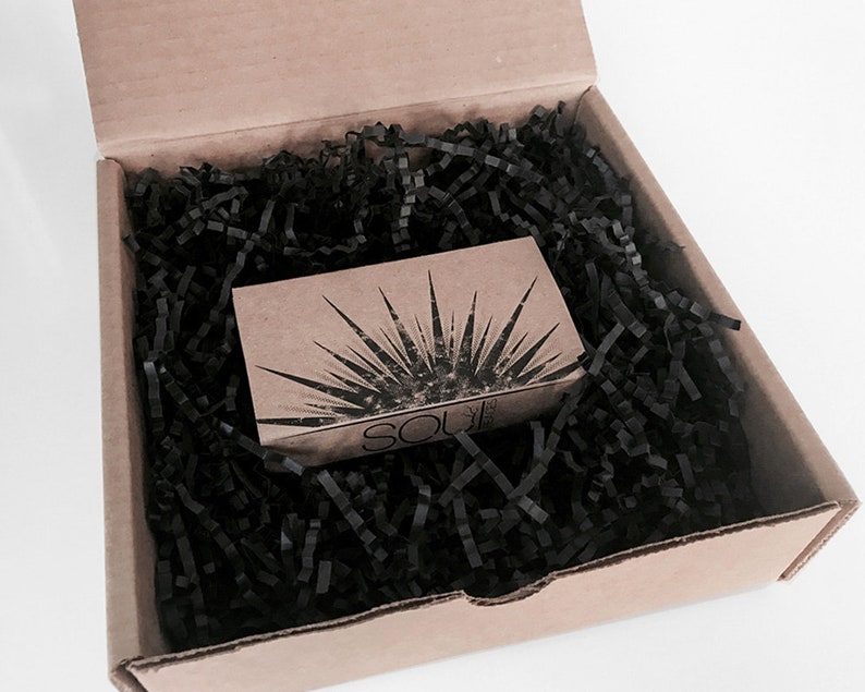 May include: A small cardboard box with black shredded paper inside. The box has a brown paper label with a sunburst design and the text "SOU" printed on it.