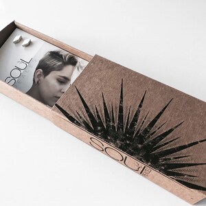May include: A small, brown cardboard box with a black and white sunburst design. The box is open and shows a white card with the text "Soul Faces" and two silver earrings.