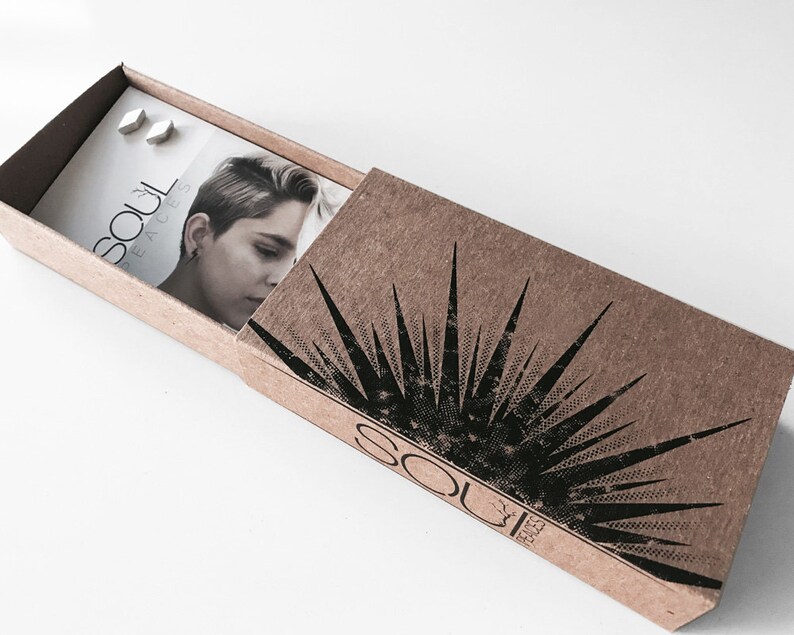 May include: A small, brown cardboard box with a black and white sunburst design. The box is open and shows a white card with the text "Soul Faces" and two silver earrings.