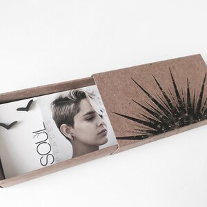 K&ouml;nnte beinhalten: Ein Paar schwarze Chevron-Ohrstecker, verpackt in einer braunen Pappbox mit einem schwarz-wei&szlig;en grafischen Design. Die Box enth&auml;lt auch eine Karte mit dem Text "Soul Peaces".