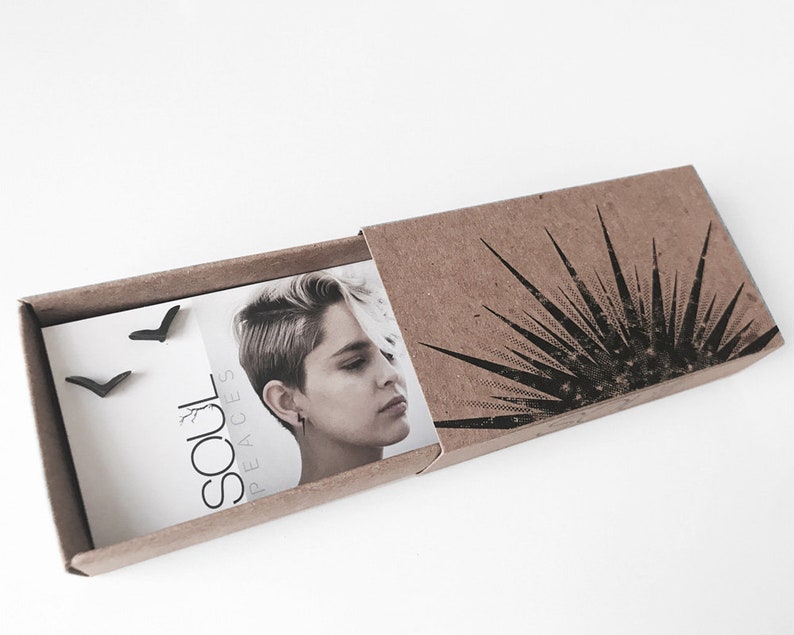 K&ouml;nnte beinhalten: Ein Paar schwarze Chevron-Ohrstecker, verpackt in einer braunen Pappbox mit einem schwarz-wei&szlig;en grafischen Design. Die Box enth&auml;lt auch eine Karte mit dem Text "Soul Peaces".