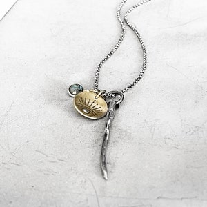 Può includere: Una collana con catena d'argento con un ciondolo dorato che presenta un designo di sole e occhio. La collana ha anche un ciondolo d'argento che assomiglia a un ramoscello.