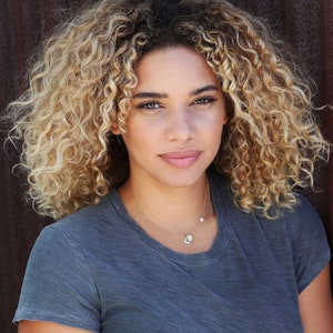May include: A woman with curly, light blonde hair wearing a gray t-shirt and a delicate gold necklace with a small star charm.