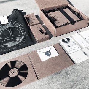 May include: A brown cardboard box with a black and white graphic design featuring a sun and moon. The box contains a black t-shirt with the text "Soul Peaces" and the phrase "live by the sun * love by the moon". The box also contains a necklace with a lightning bolt pendant, a vinyl record, and several cards with inspirational quotes.
