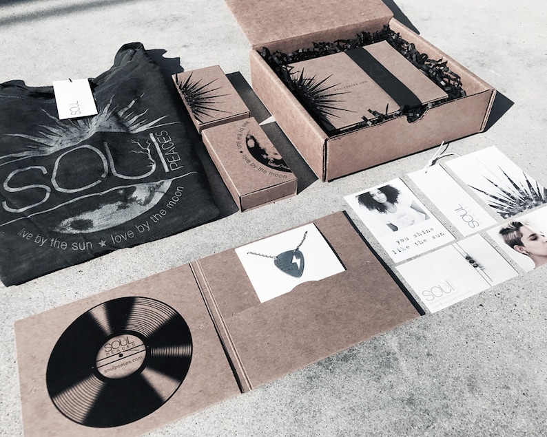 May include: A brown cardboard box with a black and white graphic design featuring a sun and moon. The box contains a black t-shirt with the text "Soul Peaces" and the phrase "live by the sun * love by the moon". The box also contains a necklace with a lightning bolt pendant, a vinyl record, and several cards with inspirational quotes.