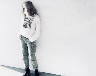 May include: A white sweatshirt with a black geometric design and the word "SOUL" printed on the front. The sweatshirt is worn by a person wearing green cargo pants and black boots.