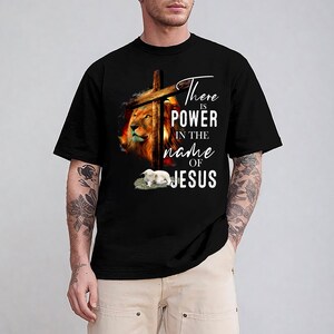 May include: Black t-shirt featuring a lion's head, a wooden cross, and a lamb. The text "There is POWER in the name of JESUS" is written in white script. The design has a religious theme.