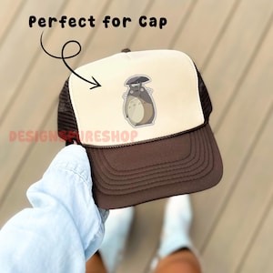 May include: A brown and tan trucker hat with a cartoon image of Totoro. The hat has a brown brim and mesh back, with the text "Perfect for Cap" above the hat. The Etsy shop name "DESIGNSFIRESHOP" is printed on the side.