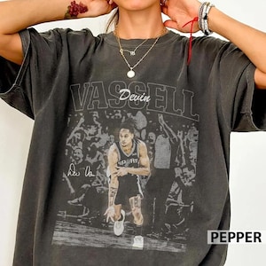 Pode incluir: Uma t-shirt cinza-carvão de manga curta com uma estampa gráfica de um jogador de basquete com uma camisola que traz o texto Devin Vassell. O design inclui um fundo de multidão desbotado e uma assinatura. Gola redonda e corte descontraído.