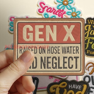 Pode incluir: Patch bordado com o texto "GEN X RAISED ON HOSE WATER AND NEGLECT" em um design retangular com borda marrom e fundo vermelho. Outros patches com desenhos florais e de script são visíveis.