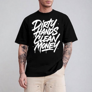 May include: Black t-shirt with the white text "Dirty Hands Clean Money" in a bold, dripping font. The shirt has a crew neck and short sleeves. The model is wearing khaki pants.