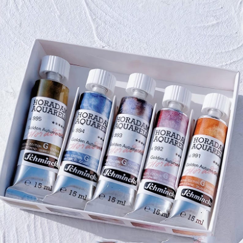 Może przedstawiać: Zestaw pięciu tubek farb akwarelowych Schmincke Horadam Aquarell o pojemności 15 ml w białym pudełku. Każda tubka ma białą zakrętkę i etykietę z napisem Horadam Aquarell, Golden Autumn oraz Schmincke, prezentującą r&oacute;żne przejścia kolor&oacute;w.