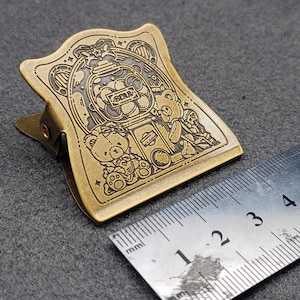 Op de afbeelding: Een gouden metalen paperclip met een gedetailleerd ontwerp van beren en een snoepgoedautomaat. Het ontwerp is gegraveerd op de paperclip en heeft een vintage uitstraling.