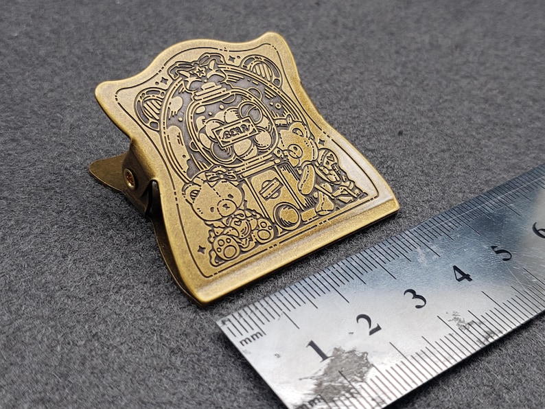Op de afbeelding: Een gouden metalen paperclip met een gedetailleerd ontwerp van beren en een snoepgoedautomaat. Het ontwerp is gegraveerd op de paperclip en heeft een vintage uitstraling.