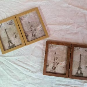 May include: Two hinged photo frames, one light wood and one dark wood, each with two panels. The panels feature images of the Eiffel Tower and the Statue of Liberty, set against a vintage map background. Decorative travel-themed frames.