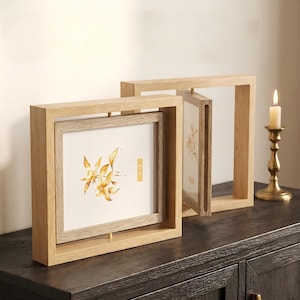 Pode incluir: Duas molduras de madeira com design quadrado. Uma moldura exibe uma impressão floral em aquarela com tons de amarelo e dourado. A outra moldura está aberta, revelando a parte de trás da impressão. Uma vela acesa está à direita.