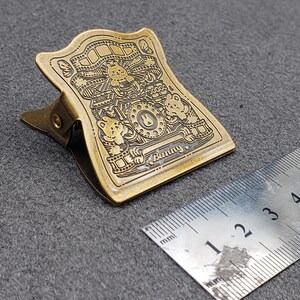 Op de afbeelding: Een gouden metalen binderclip met een ingewikkeld ontwerp met cartoondieren en het woord "Bunny".