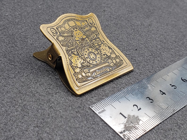 Op de afbeelding: Een gouden metalen binderclip met een ingewikkeld ontwerp met cartoondieren en het woord "Bunny".