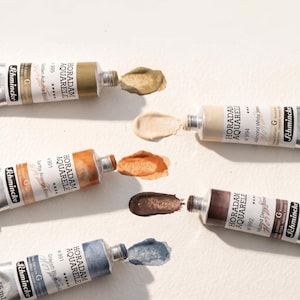 Könnte beinhalten: Nahaufnahme von sechs Tuben Schmincke Horadam Aquarell Aquarellfarben. Die silbernen Tuben haben verschiedene Farben, darunter Brauntöne, Beige und Blau. Der Text auf den Tuben ist sichtbar.