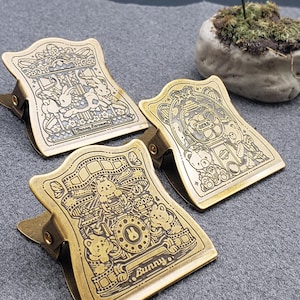 Op de afbeelding: Drie gouden metalen binderclips met verschillende ontwerpen. De eerste clip heeft een carrousel met dieren, de tweede clip heeft een beer in een automaat en de derde clip heeft een konijn in een telefooncel. De derde clip heeft het woord "Bunny" erop gedrukt.