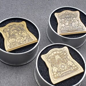 Op de afbeelding: Drie gouden metalen pin badges met verschillende ontwerpen met cartoonberen en het woord "Sunny". De badges worden getoond in ronde, zilverkleurige containers met zwarte schuimrubberen inzetstukken.