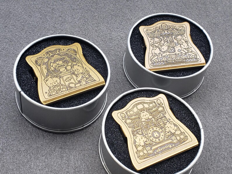 Op de afbeelding: Drie gouden metalen pin badges met verschillende ontwerpen met cartoonberen en het woord "Sunny". De badges worden getoond in ronde, zilverkleurige containers met zwarte schuimrubberen inzetstukken.