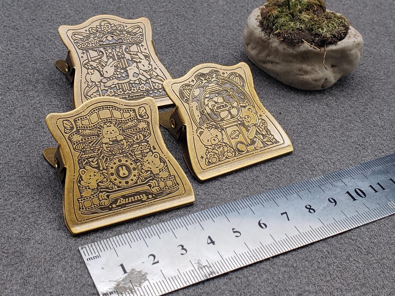 Op de afbeelding: Drie gouden metalen binderclips met verschillende cartoon-dierontwerpen. De clips tonen een draaimolen, een beer in een automaat en een konijn in een fotohokje. De tekst "Bunny." staat onderaan de konijnenclip.