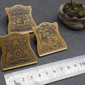 Op de afbeelding: Drie gouden metalen binderclips met ingewikkelde ontwerpen met cartoondieren en het woord "Bunny".