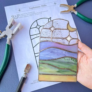 Könnte beinhalten: Ein Buntglasfenster, das eine nächtliche Landschaft mit Hügeln, einer Mondsichel und Sternen darstellt. Das Glas ist in kleine Stücke geschnitten und in einem Rahmen zusammengefügt. Das Stück ist etwa 10x17 Zoll groß.