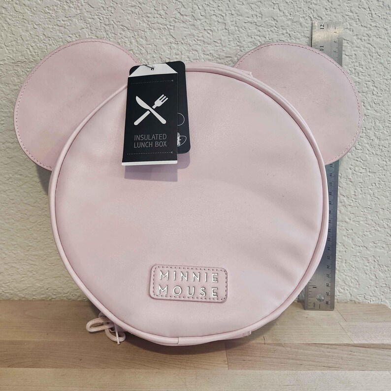 May include: A light pink, round insulated lunch box with attached ears, resembling a cartoon character. The lunch box has a tag that reads "INSULATED LUNCH BOX" and a "MINNIE MOUSE" label. A ruler is placed next to the lunch box.