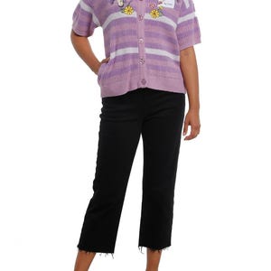 May include: A short-sleeved, button-up cardigan in lavender and white stripes. The sweater features embroidered floral and cartoon designs. The person is wearing black cropped jeans and black flats.