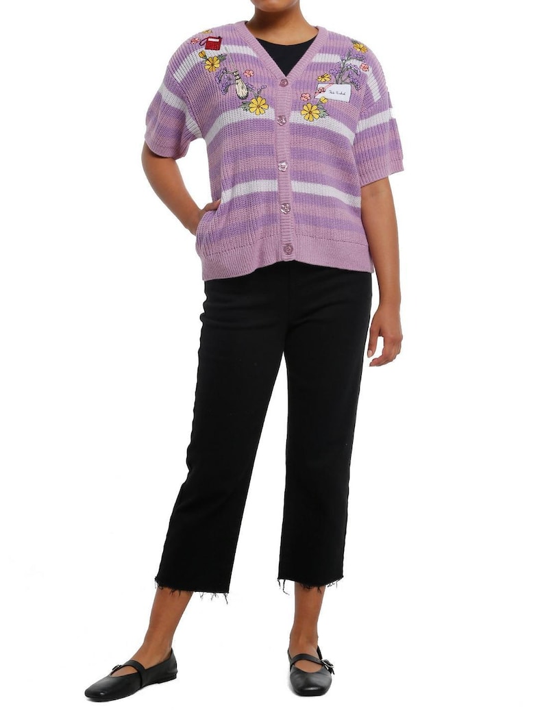 May include: A short-sleeved, button-up cardigan in lavender and white stripes. The sweater features embroidered floral and cartoon designs. The person is wearing black cropped jeans and black flats.
