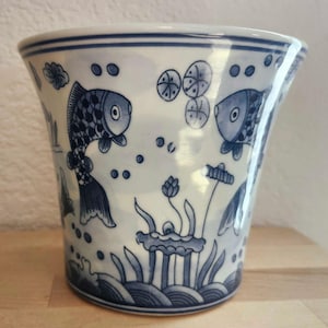 May include: A white and blue ceramic vase with a flared rim. The vase features a hand-painted design of fish, aquatic plants, and bubbles. The fish are detailed with scales and fins, and the plants include lotus flowers.