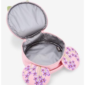 May include: Pink lunch bag with a green handle and two round side pockets. The side pockets and the bag's front feature a purple floral design. The interior is gray and insulated. The bag is open, revealing the inside.