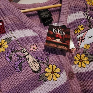 May include: A purple cardigan sweater with white stripes and embroidered floral and broomstick details. The sweater features a "Kiki's Delivery Service" tag. The design includes yellow flowers, purple flowers, and a red mailbox.