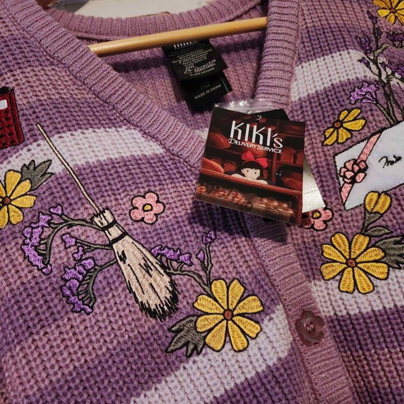 May include: A purple cardigan sweater with white stripes and embroidered floral and broomstick details. The sweater features a "Kiki's Delivery Service" tag. The design includes yellow flowers, purple flowers, and a red mailbox.