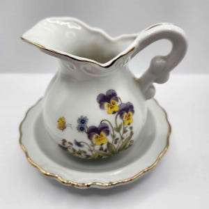 May include: White ceramic pitcher and saucer set. The pitcher features a floral design with purple and yellow pansies, a small butterfly, and gold trim. The saucer has a scalloped edge and gold trim. The handle is curved.