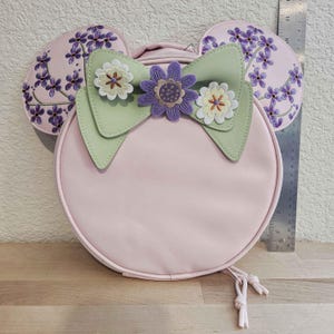 May include: A light pink, round backpack with attached mouse ears. The ears are decorated with purple floral embroidery. A light green bow with flower embellishments adorns the front. The backpack has a drawstring closure.