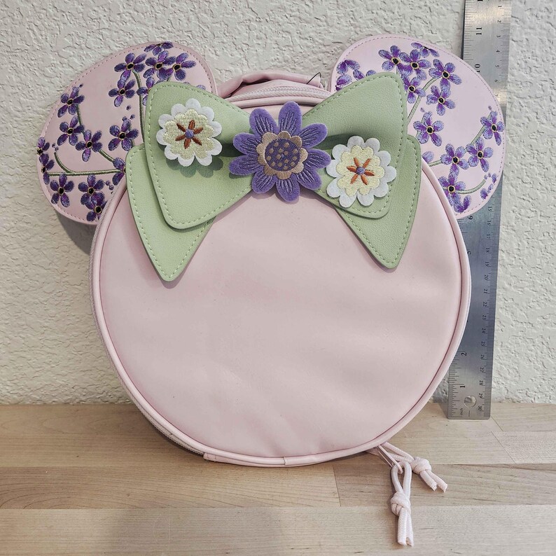 May include: A light pink, round backpack with attached mouse ears. The ears are decorated with purple floral embroidery. A light green bow with flower embellishments adorns the front. The backpack has a drawstring closure.