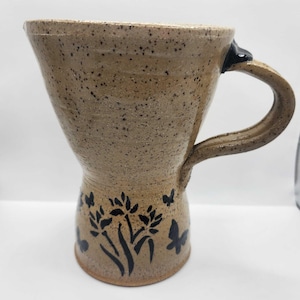 May include: A handmade ceramic mug with a speckled tan finish. The mug features a flared top and a curved handle. Black floral and butterfly designs decorate the lower portion of the mug. The mug is approximately 5 inches tall.