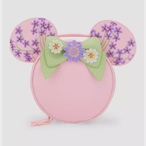 May include: A pink round handbag with attached mouse ears. The ears are decorated with purple floral patterns. A green bow with white and purple flower accents adorns the front. The bag has a top handle and a zipper closure.