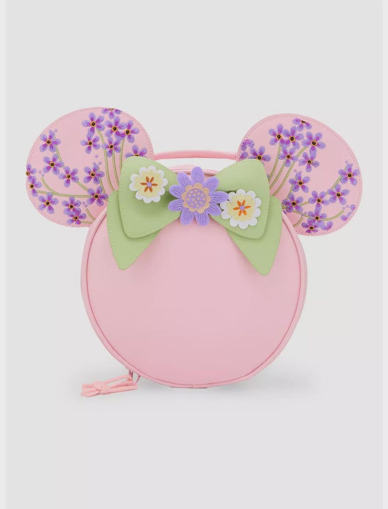 May include: A pink round handbag with attached mouse ears. The ears are decorated with purple floral patterns. A green bow with white and purple flower accents adorns the front. The bag has a top handle and a zipper closure.