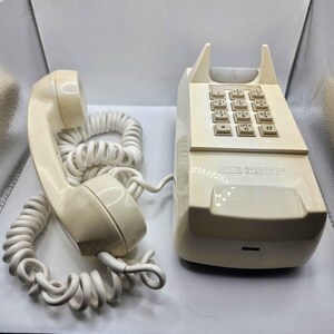 May include: Ivory-colored vintage telephone with a handset and coiled cord. The base features a keypad with number and letter keys, and the text "GTE Automatic Electric". A classic, retro communication device.