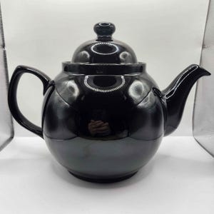 May include: A glossy black teapot with a matching lid and handle. The teapot has a rounded body and a curved spout. The lid is topped with a small, round knob. The teapot is designed for brewing tea.